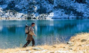 Man hiking with a backpack beside a mountain lake, carrying a fishing rod.