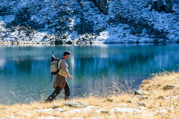 Man hiking with a backpack beside a mountain lake, carrying a fishing rod.