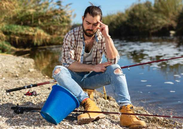Frustrated man sitting by a river, head in hand, near his fishing rod.