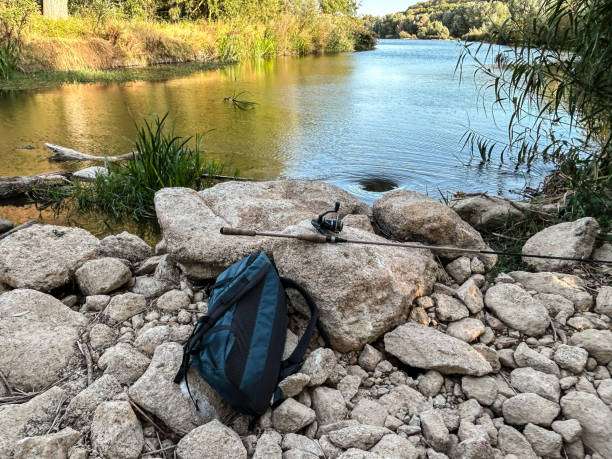 Fishing rod and backpack resting on rocks beside a calm river, ready for use.