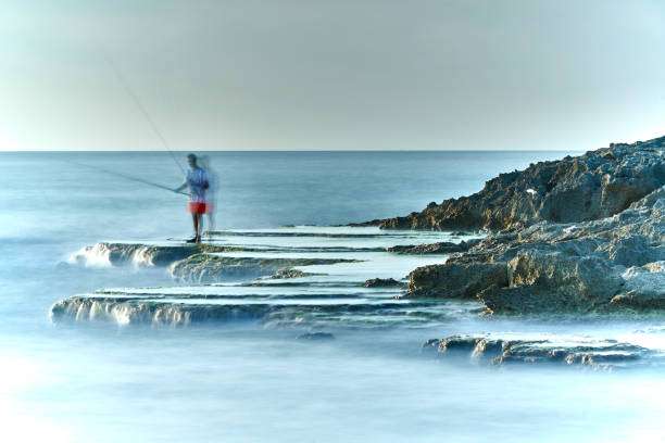 Fisherman on rocky coast with fishing reel, long exposure sea.